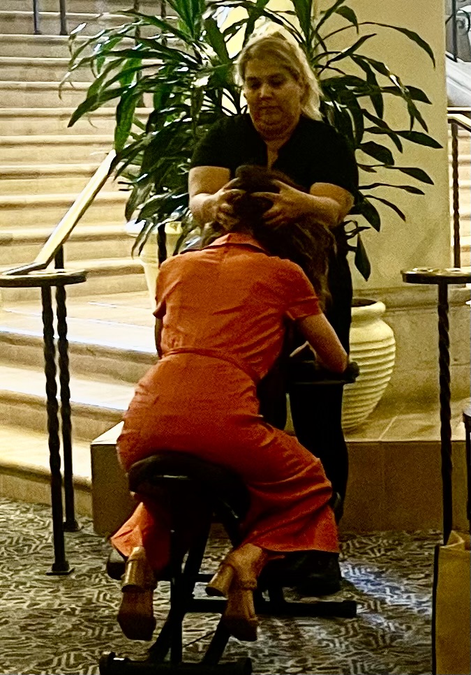 A woman relaxes in a chair as her hair is gently combed, embodying tranquility amidst a busy corporate event.